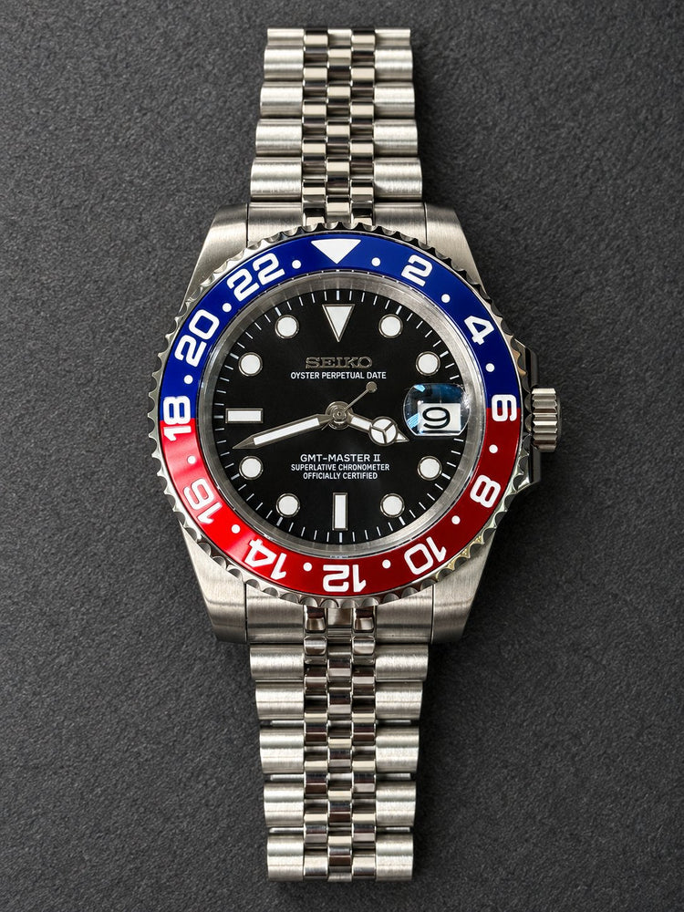 Front view of a custom Seiko 3-hand automatic watch with black dial, blue-and-red 24-hour bezel and steel jubilee-style bracelet