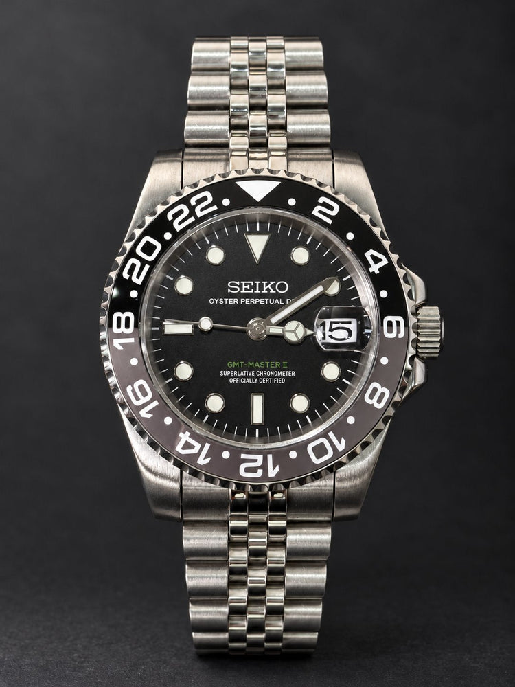 Front view of a custom Seiko 3-hand automatic watch with black dial, black-and-grey 24-hour bezel and steel jubilee-style bracelet